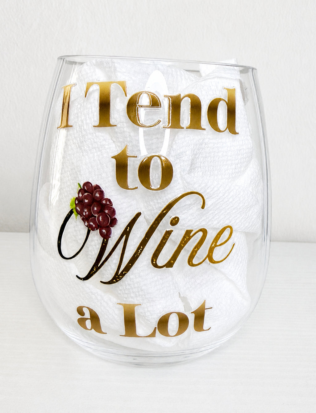 A clear shatterproof stemless wine glass with the phrase 'I Tend to Wine a Lot' in metallic gold lettering, featuring a small red grape graphic, set against a white background.