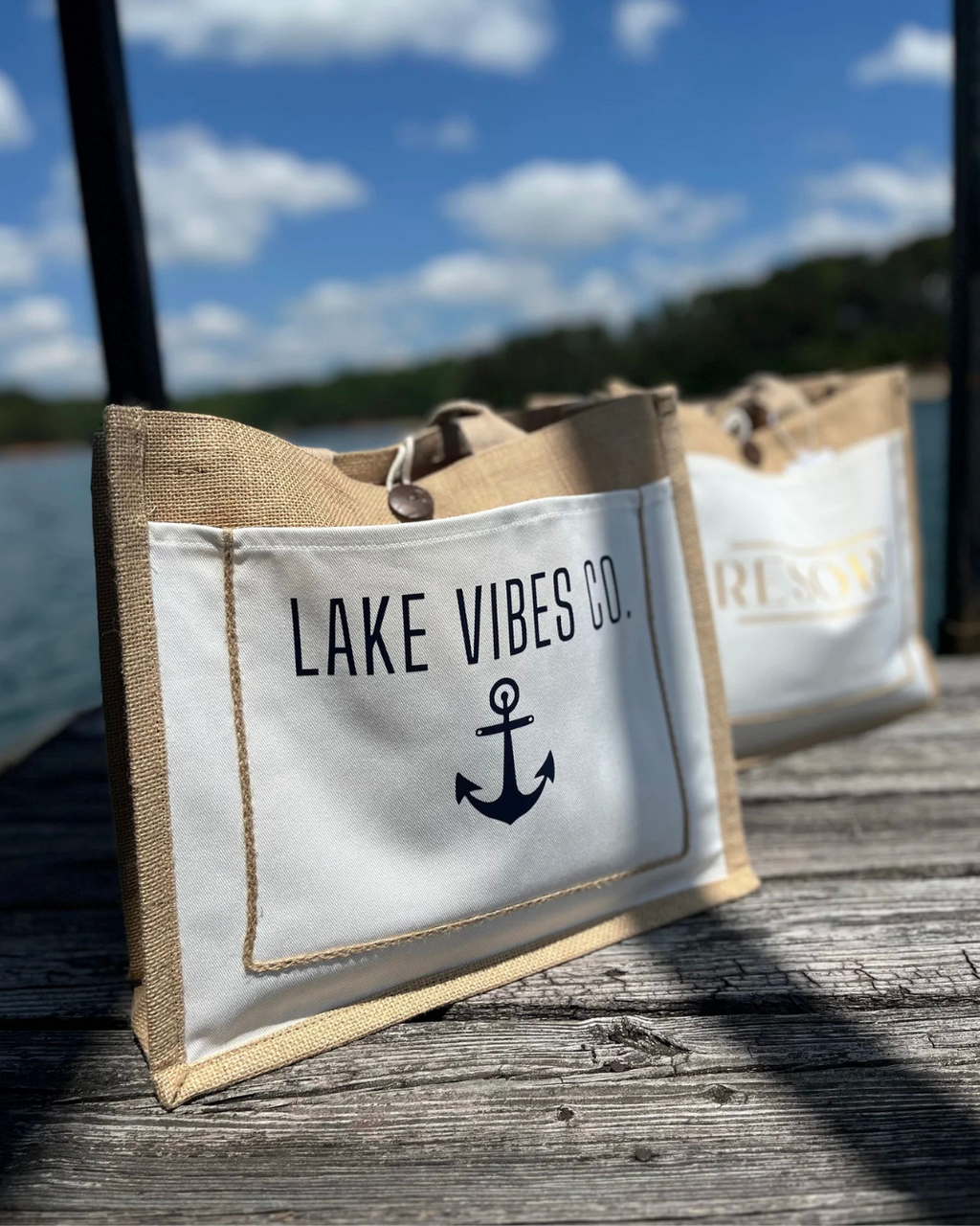 A canvas tote bag with a natural burlap-style top and bottom and a white front panel printed with the text 'LAKE VIBES CO.' and a dark anchor graphic, photographed on a wooden dock with water and trees in the background.