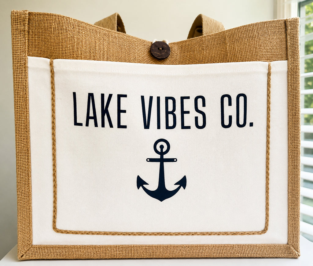 A burlap and canvas tote bag with a white front panel printed with the text 'LAKE VIBES CO.' and a navy anchor graphic.