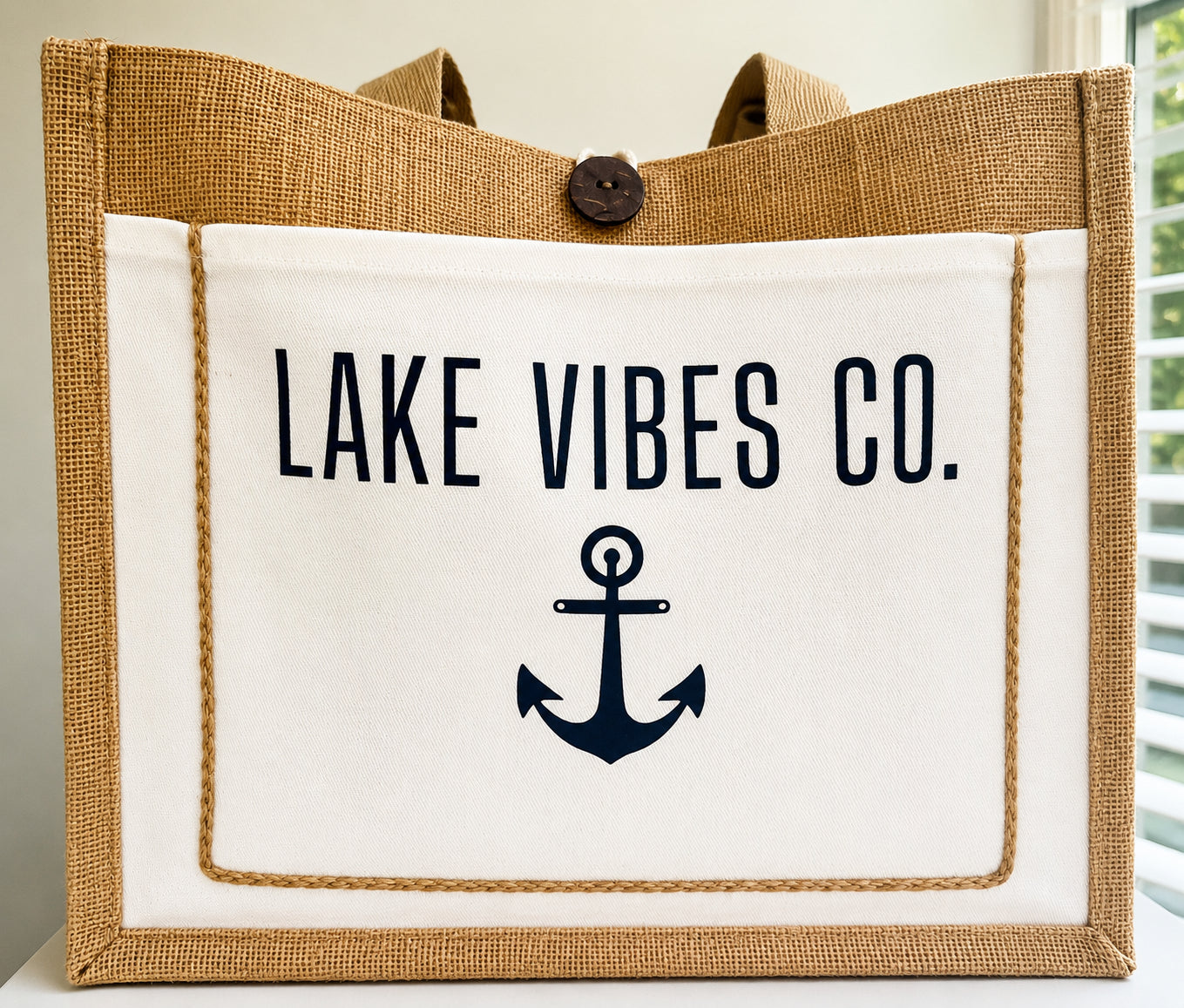 A burlap and canvas tote bag with a white front panel printed with the text 'LAKE VIBES CO.' and a navy anchor graphic.