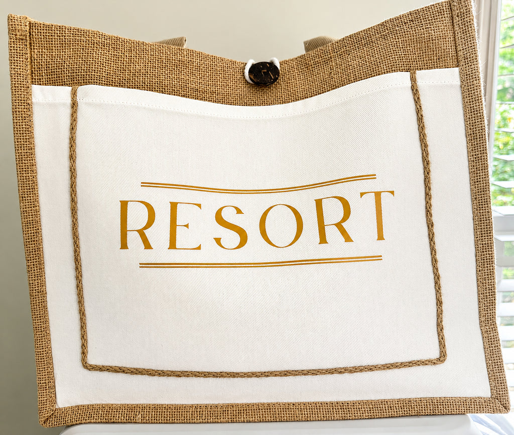 A tote bag with a white front panel printed with the word 'RESORT' in gold, a natural burlap border, and a black button closure at the top, shown against an indoor background with a window visible on the right.