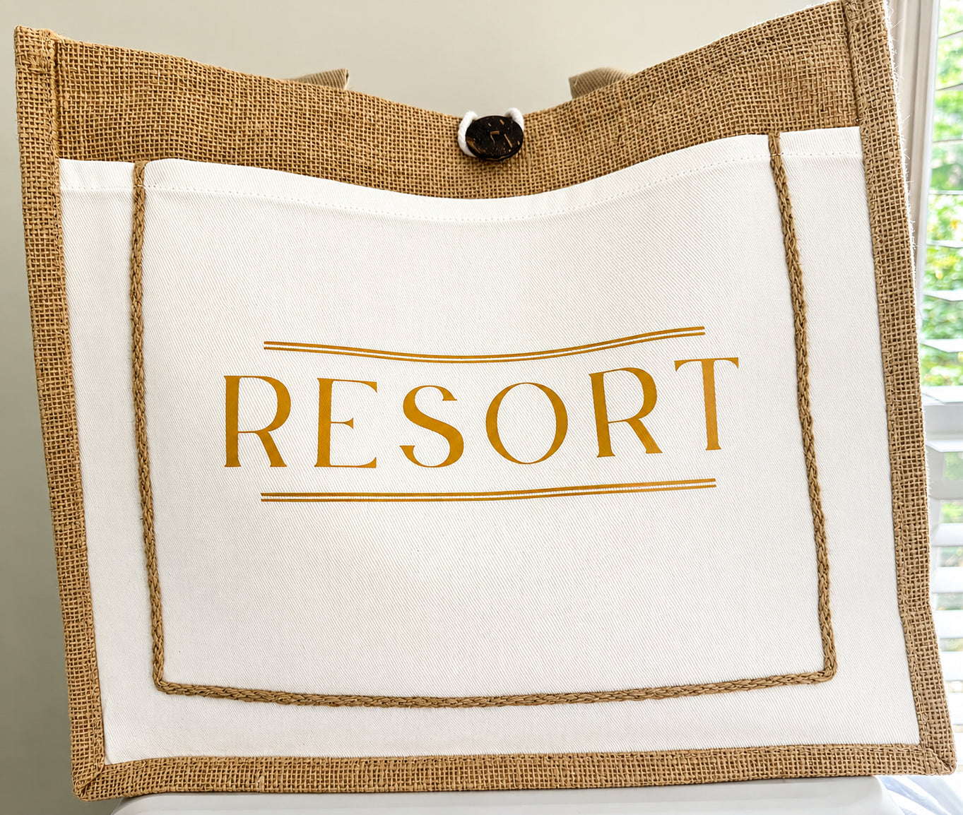 A tote bag with a white front panel printed with the word 'RESORT' in gold, a natural burlap border, and a black button closure at the top, shown against an indoor background with a window visible on the right.