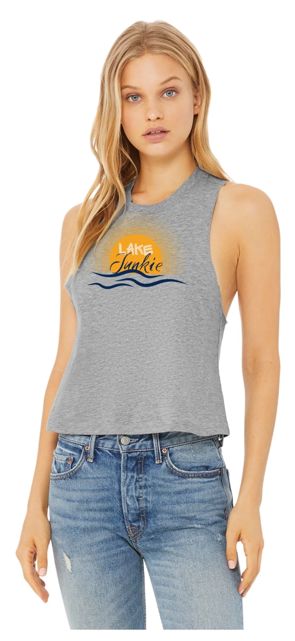A woman modeling a heather gray sleeveless crop tank top with a 'Lake Junkie' sunset graphic on the chest, paired with blue jeans against a plain white background.
