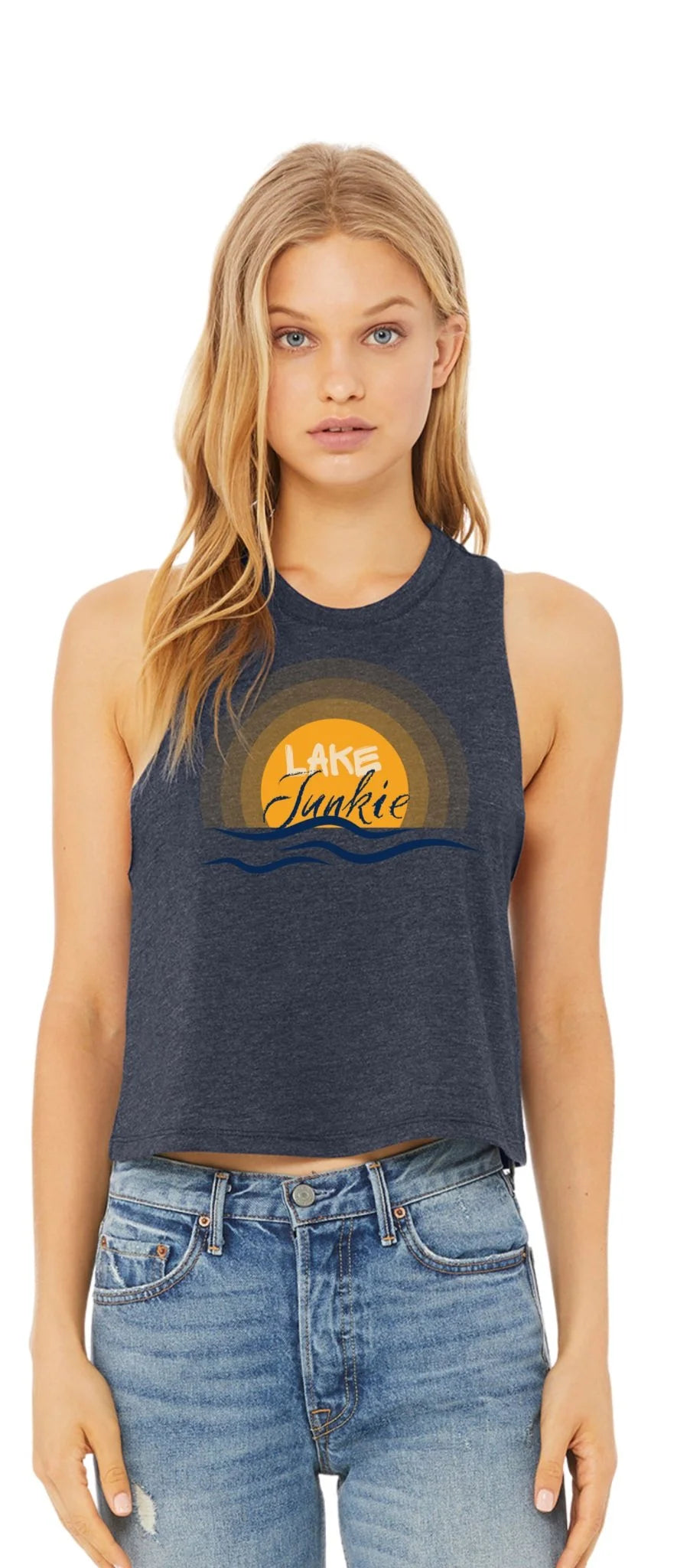 A woman modeling a dark heather gray sleeveless crop top with a 'Lake Junkie' sunset graphic in yellow and orange, paired with blue jeans, against a plain white background.