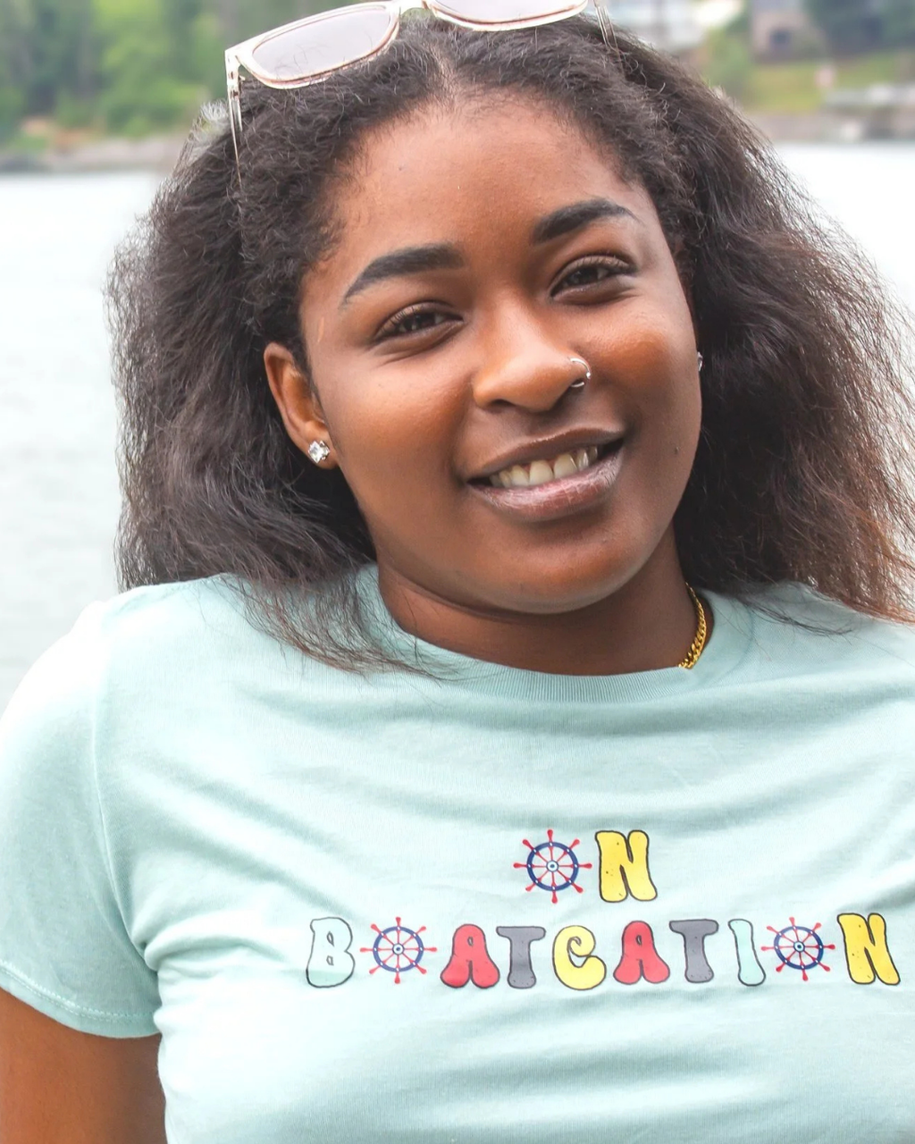 A smiling woman with dark curly hair wearing a light blue t-shirt with 'ON BOATATION' printed in colorful letters, with sunglasses on her head and a lake in the background.