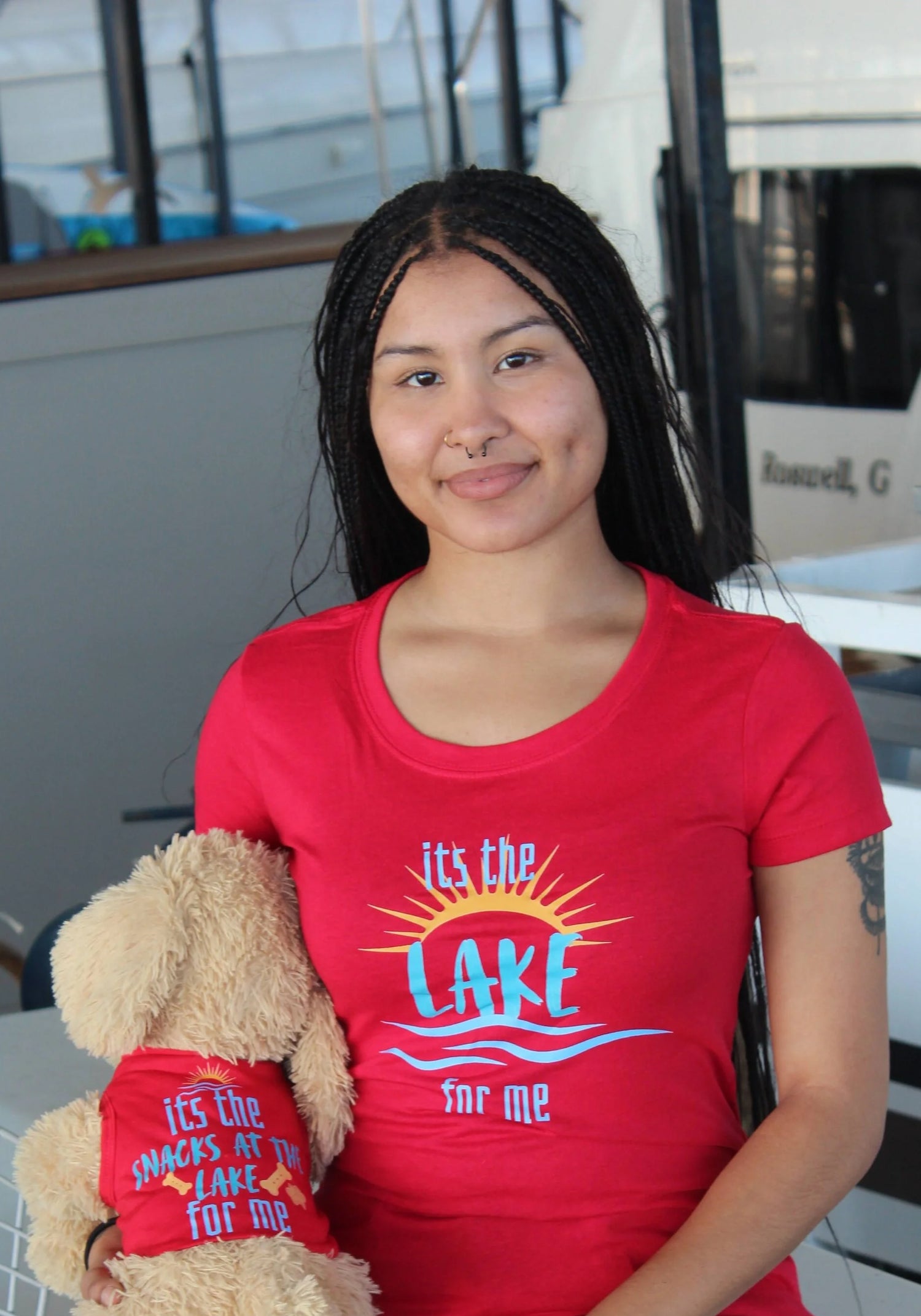A woman wearing a bright red graphic t-shirt with the text 'its the LAKE for me' in blue and yellow, holding a plush dog wearing a matching red shirt that says "it's the snacks at the lake for me"