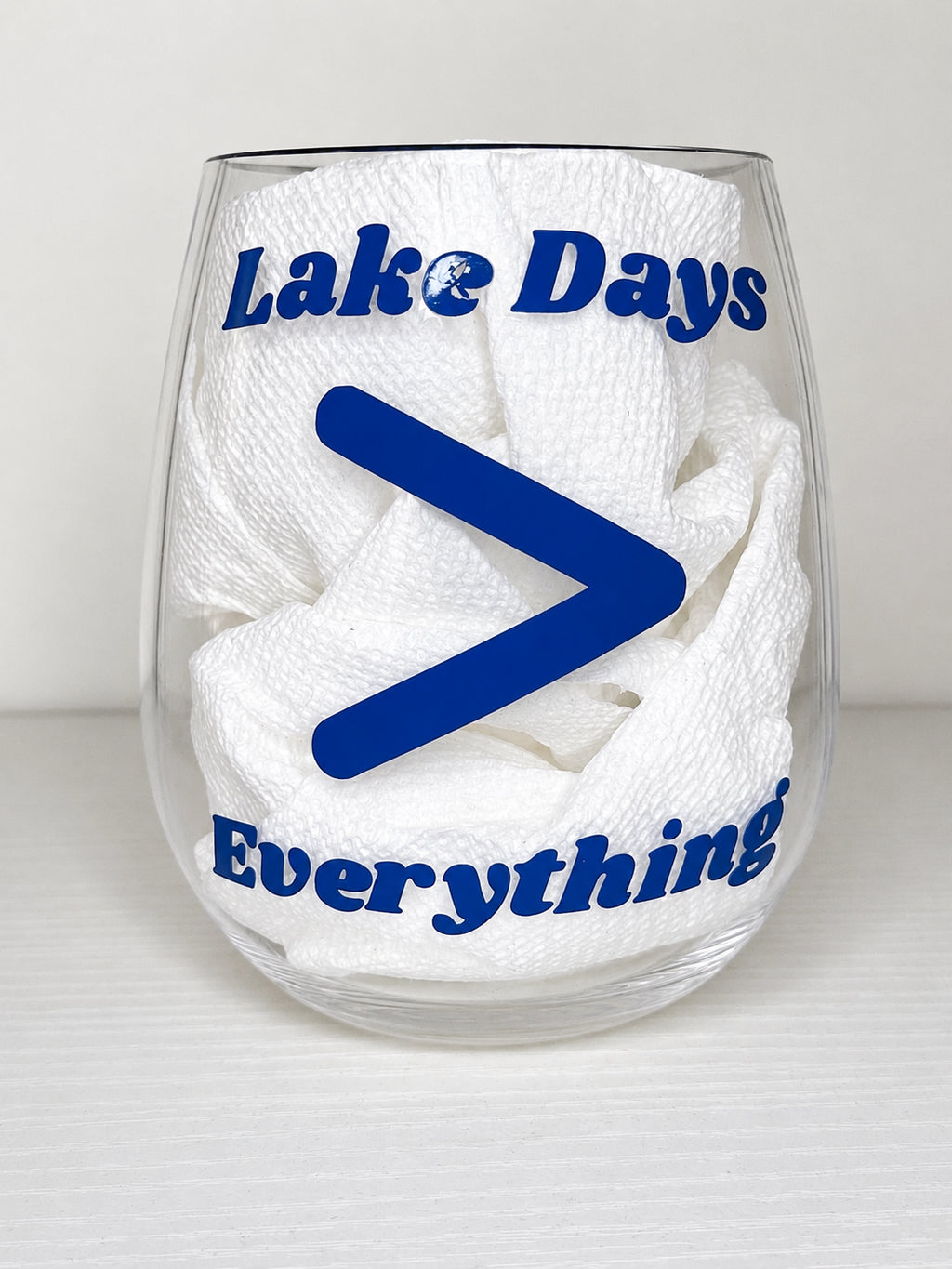 A clear stemless wine glass with blue text reading 'Lake Days Everything' and a large blue greater-than symbol, on a light gray background.