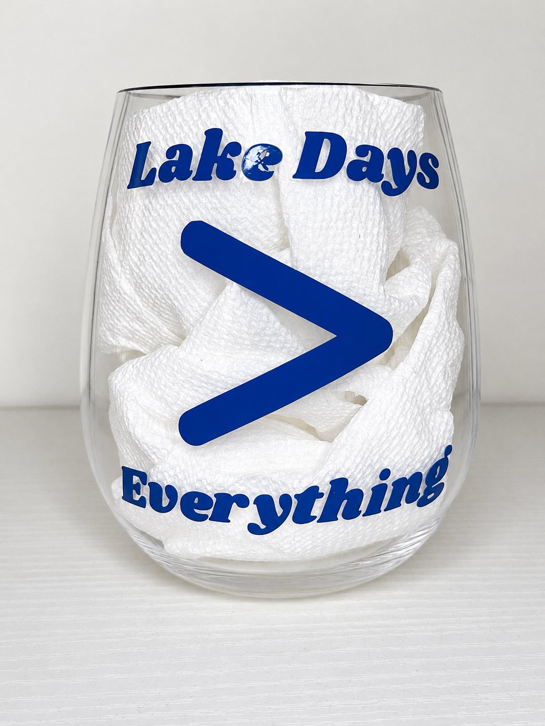 A clear stemless wine glass with blue text reading 'Lake Days Everything' and a large blue greater-than symbol, on a light gray background.