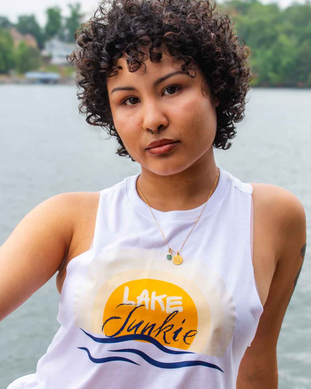 A woman with curly dark hair wearing a white tank top with a 'Lake Junkie' graphic, photographed outdoors with water and trees in the background.