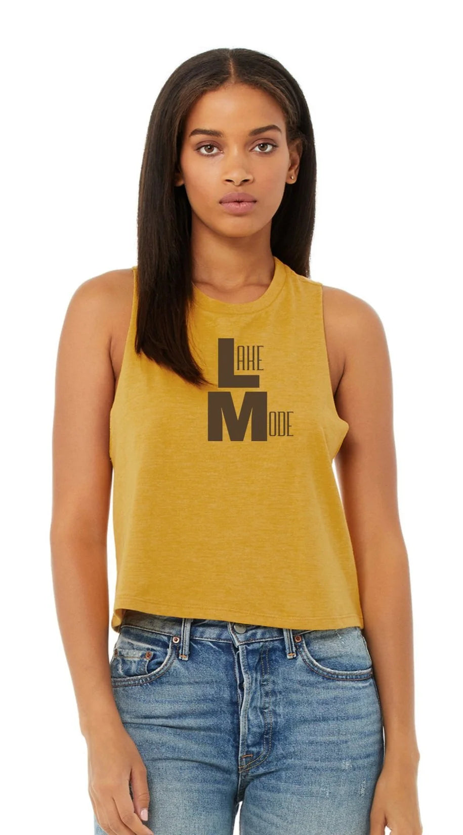 A woman modeling a mustard yellow sleeveless crop top with a black 'LAKE MODE' graphic on the front, paired with blue jeans against a plain white background.