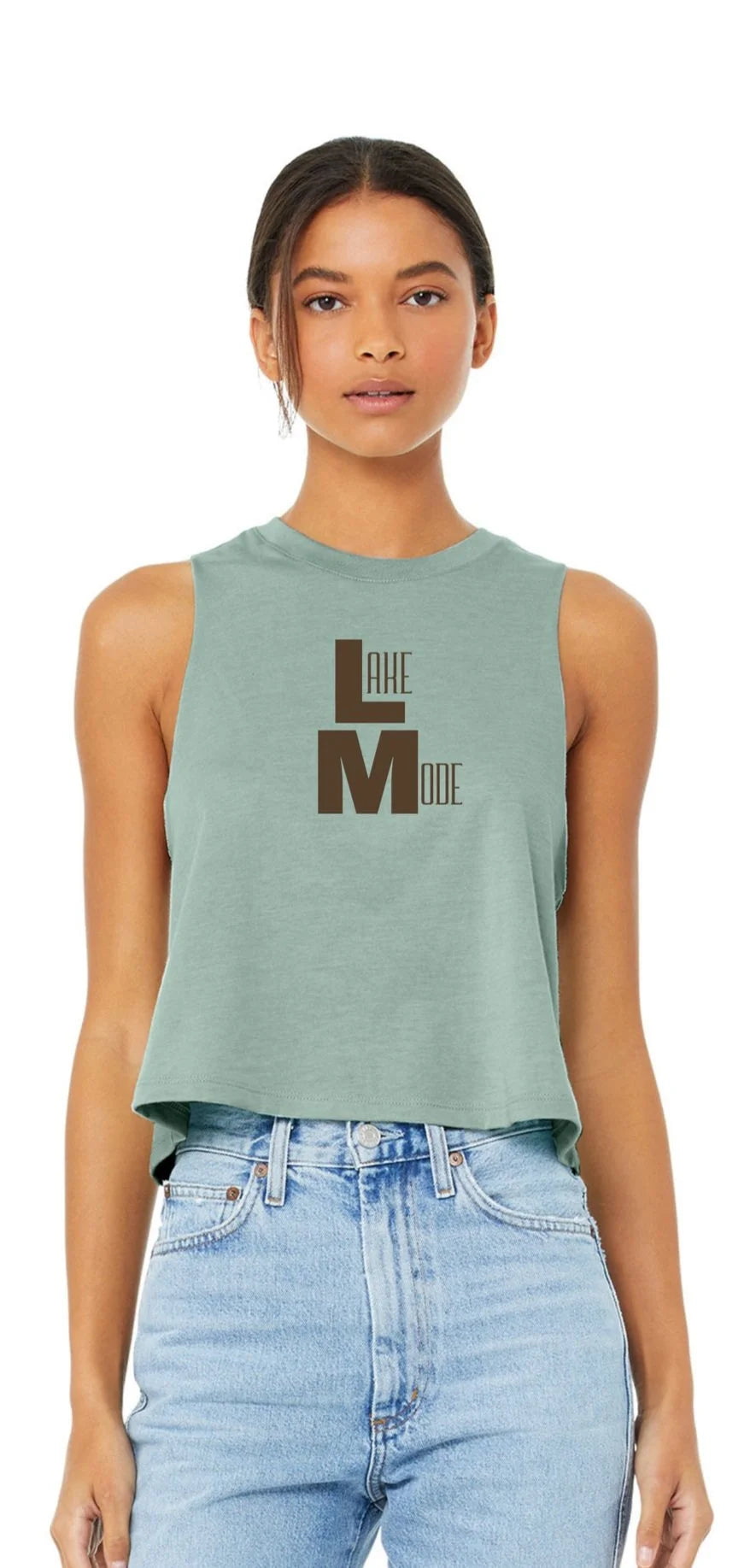 A woman modeling a sleeveless cropped tank top in sage green with a dark brown graphic reading LAKE MODE on the front, paired with light blue jeans against a plain white background.