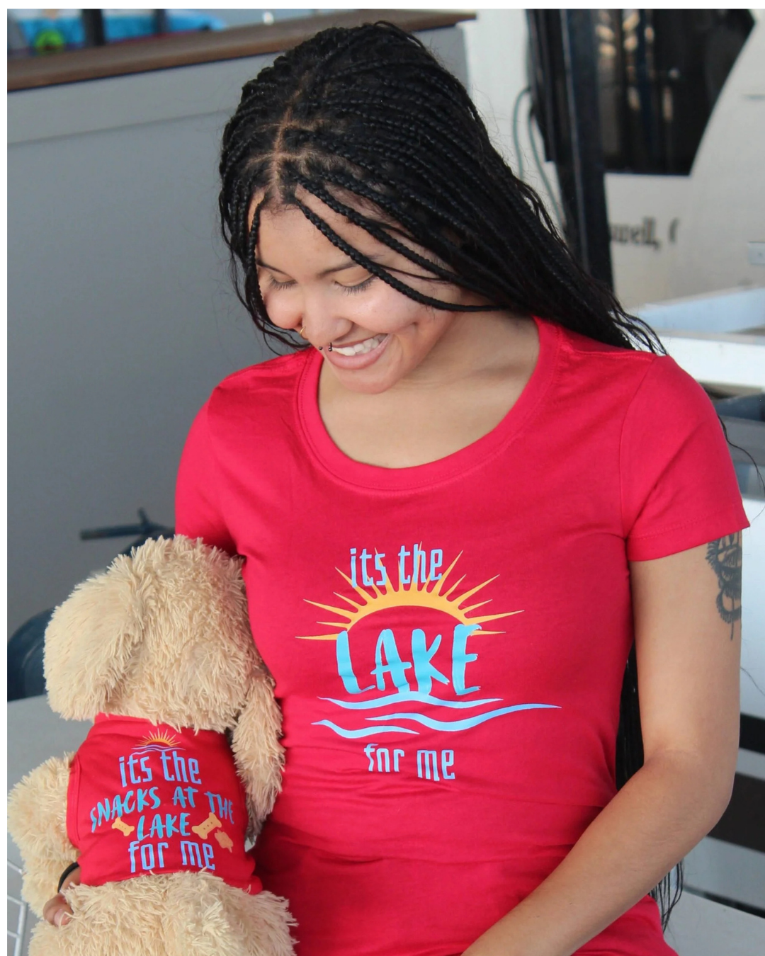 A woman wearing a bright red graphic t-shirt with the text 'its the LAKE for me' in blue and yellow, holding a plush dog wearing a matching red shirt that says "it's the snacks at the lake for me"