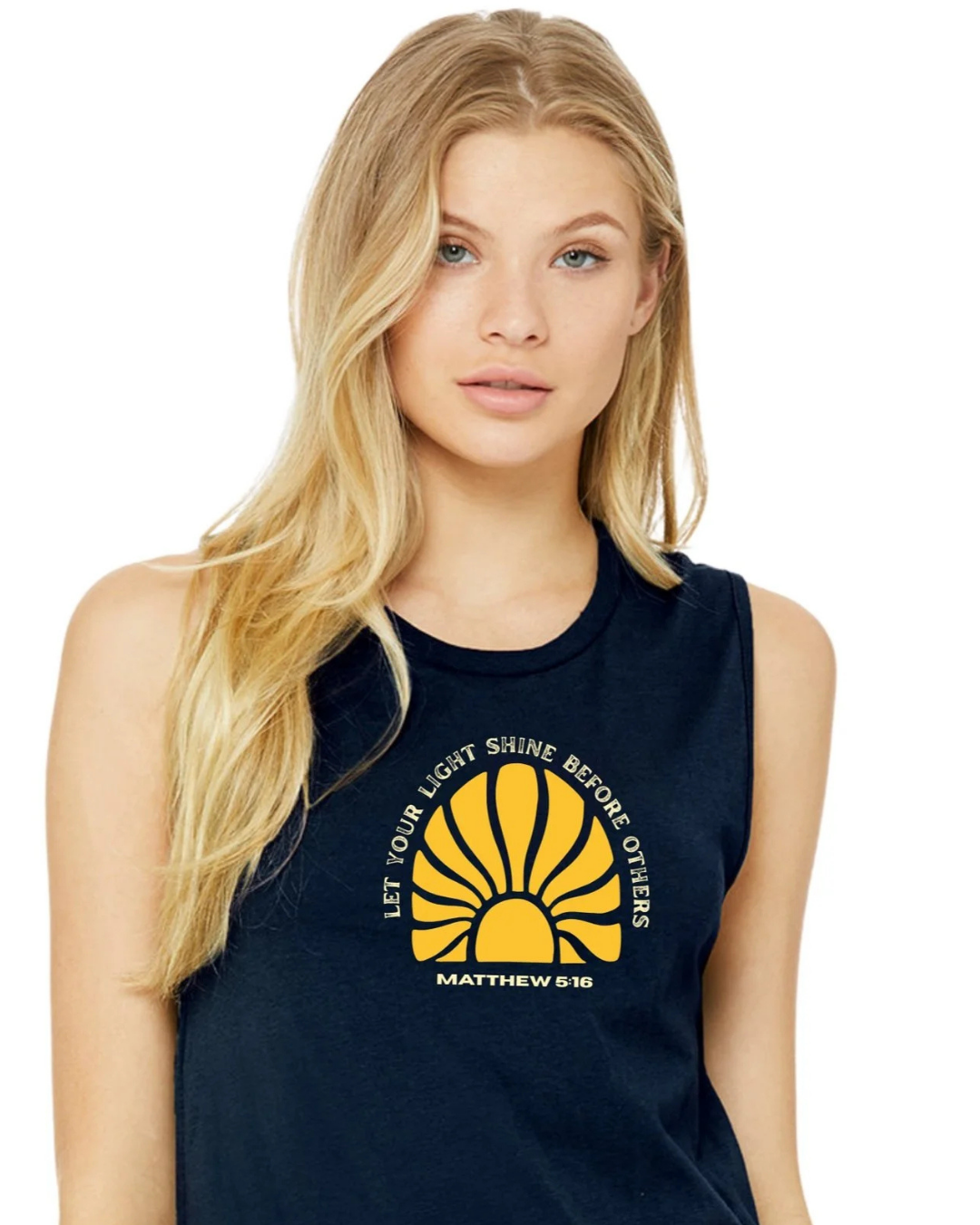 A blonde woman wearing a navy blue sleeveless crop top with a yellow sun graphic and white words that read "Let your light shine before others" Matthew 5:16 graphic on the front, set against a plain white background.