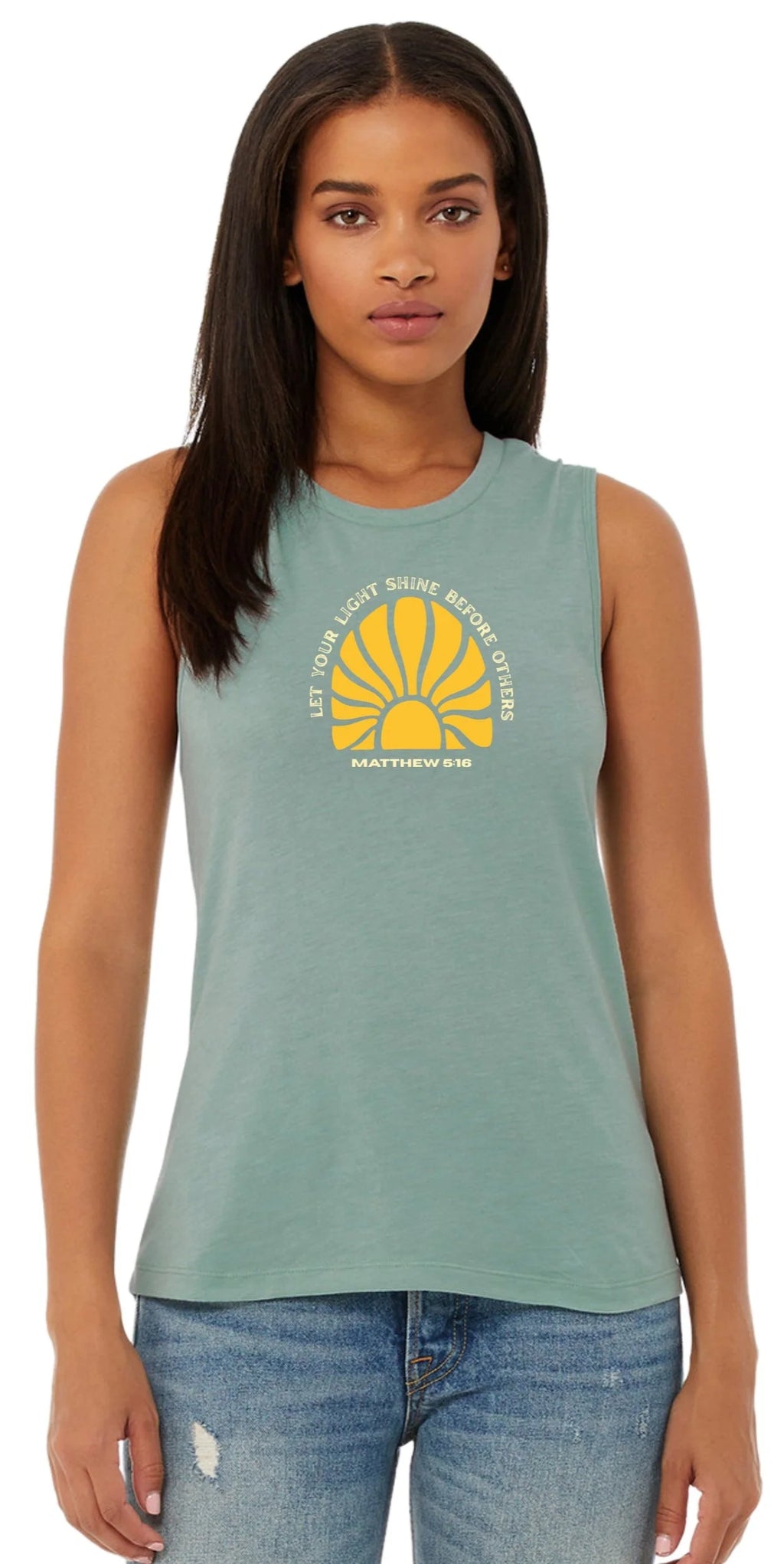 A woman modeling a sleeveless crewneck tank top in a dusty sage green color, featuring a yellow and white graphic on the chest with a sunburst and the text 'LET YOUR LIGHT shine BEFORE OTHERS' and 'MATTHEW 5:16'. She is wearing blue jeans against a plain white background.