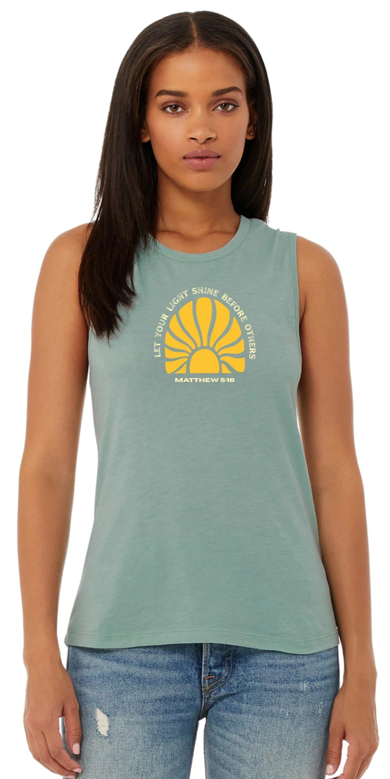 A woman modeling a sleeveless crewneck tank top in a dusty sage green color, featuring a yellow and white graphic on the chest with a sunburst and the text 'LET YOUR LIGHT shine BEFORE OTHERS' and 'MATTHEW 5:16'. She is wearing blue jeans against a plain white background.