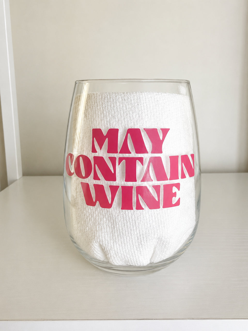 A clear shatterproof stemless wine glass with bold pink text reading 'MAY CONTAIN WINE' on the front, set on a light surface against a neutral background.
