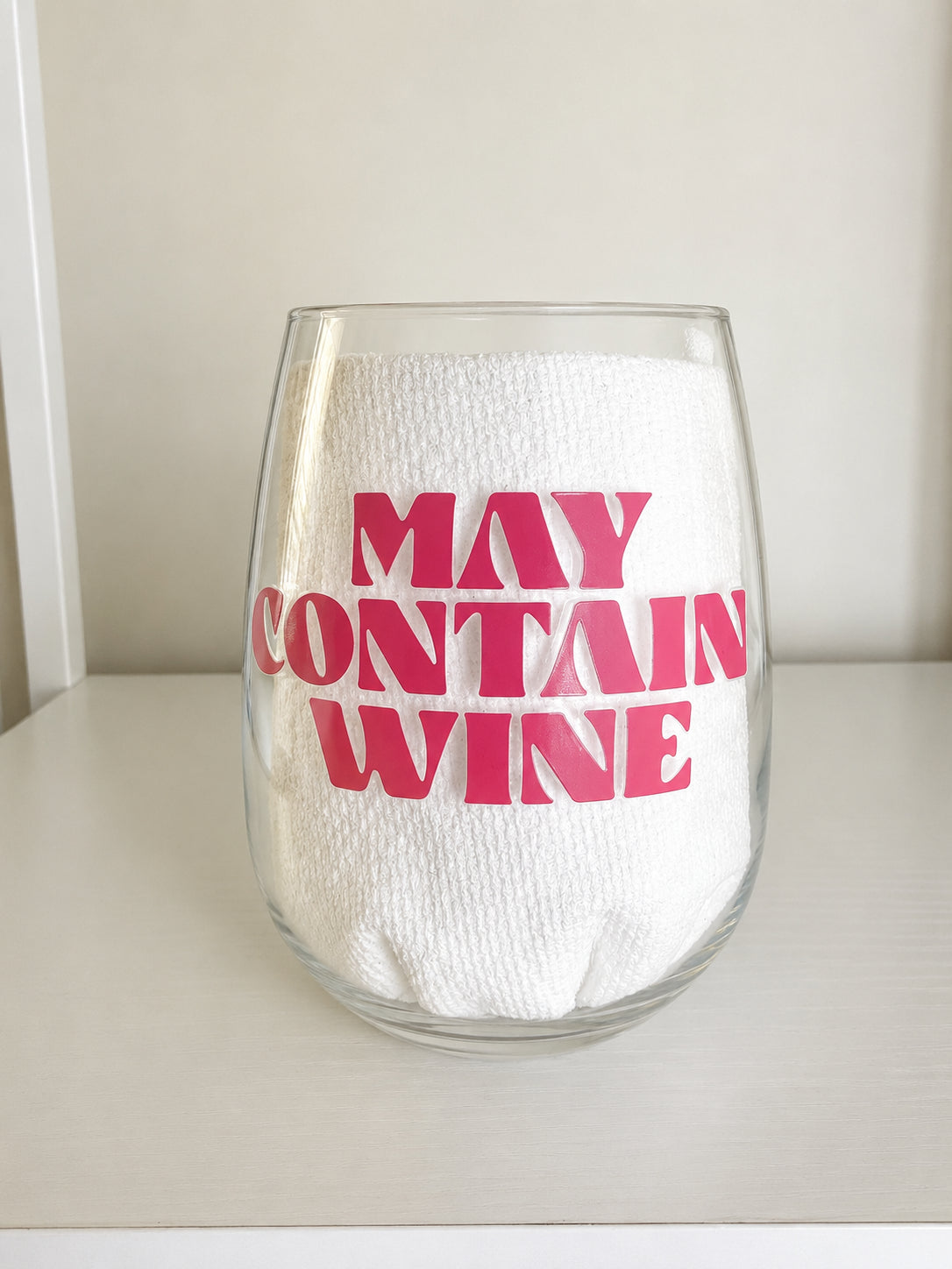 A clear shatterproof stemless wine glass with bold pink text reading 'MAY CONTAIN WINE' on the front, set on a light surface against a neutral background.