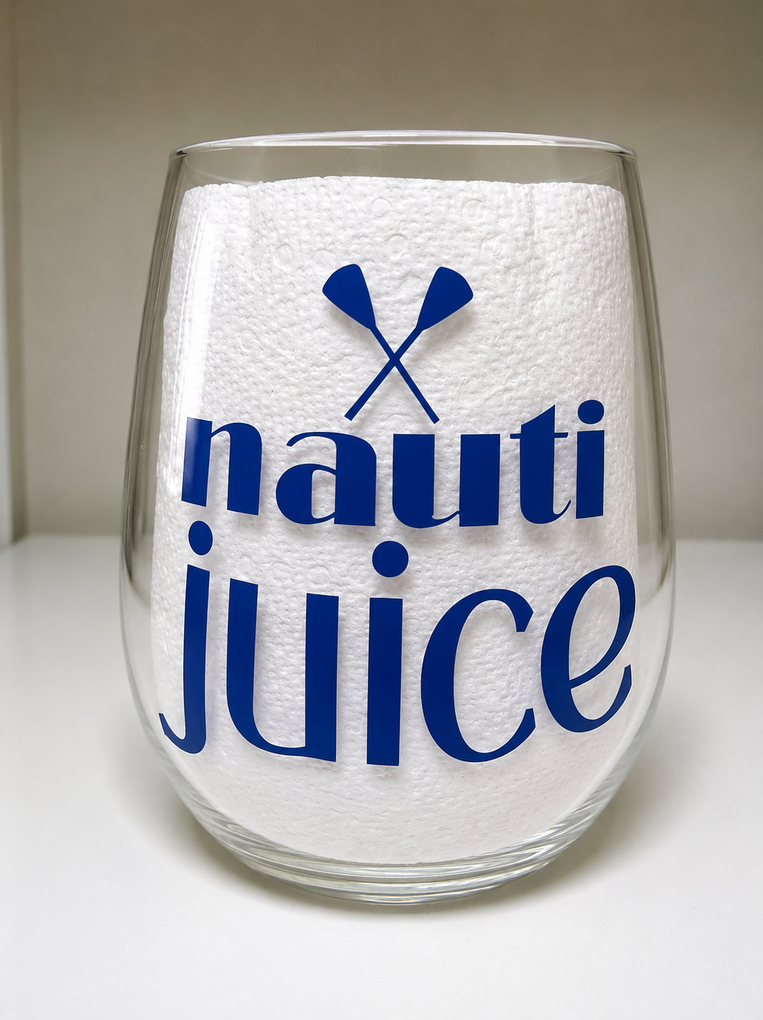 A clear shatterproof stemless wine glass, printed with the words 'nauti juice' in dark blue and crossed oars above it, shown against a neutral beige and white background.
