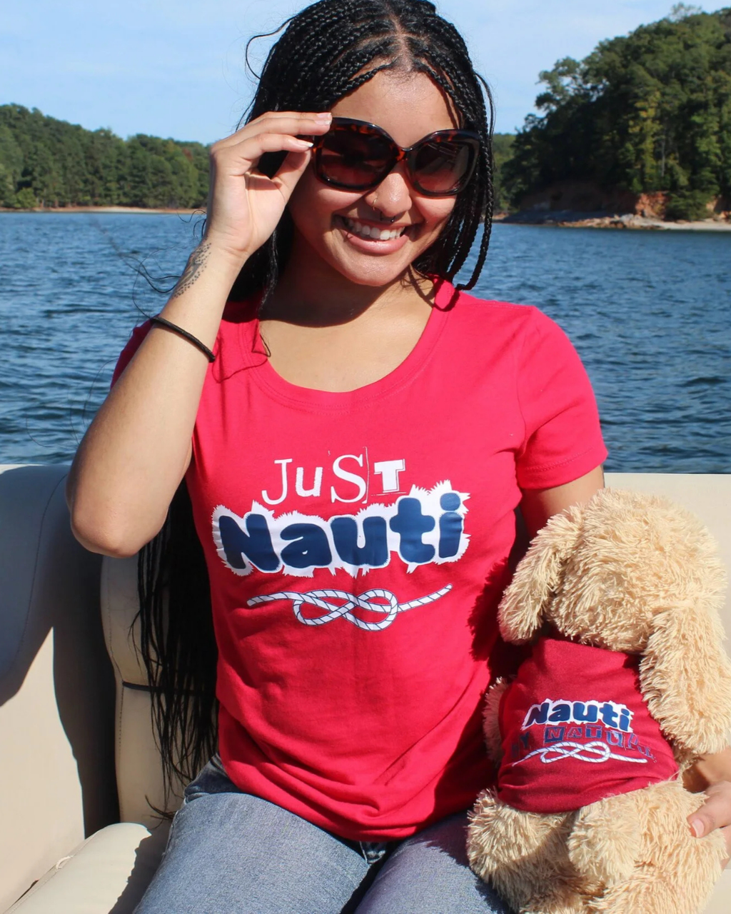 A woman sitting on a boat wearing a bright red graphic t-shirt with 'Just Nauti' printed in white and blue, holding a fluffy dog that matches the shirt design. The background shows blue water and a tree-lined shore under a clear sky.