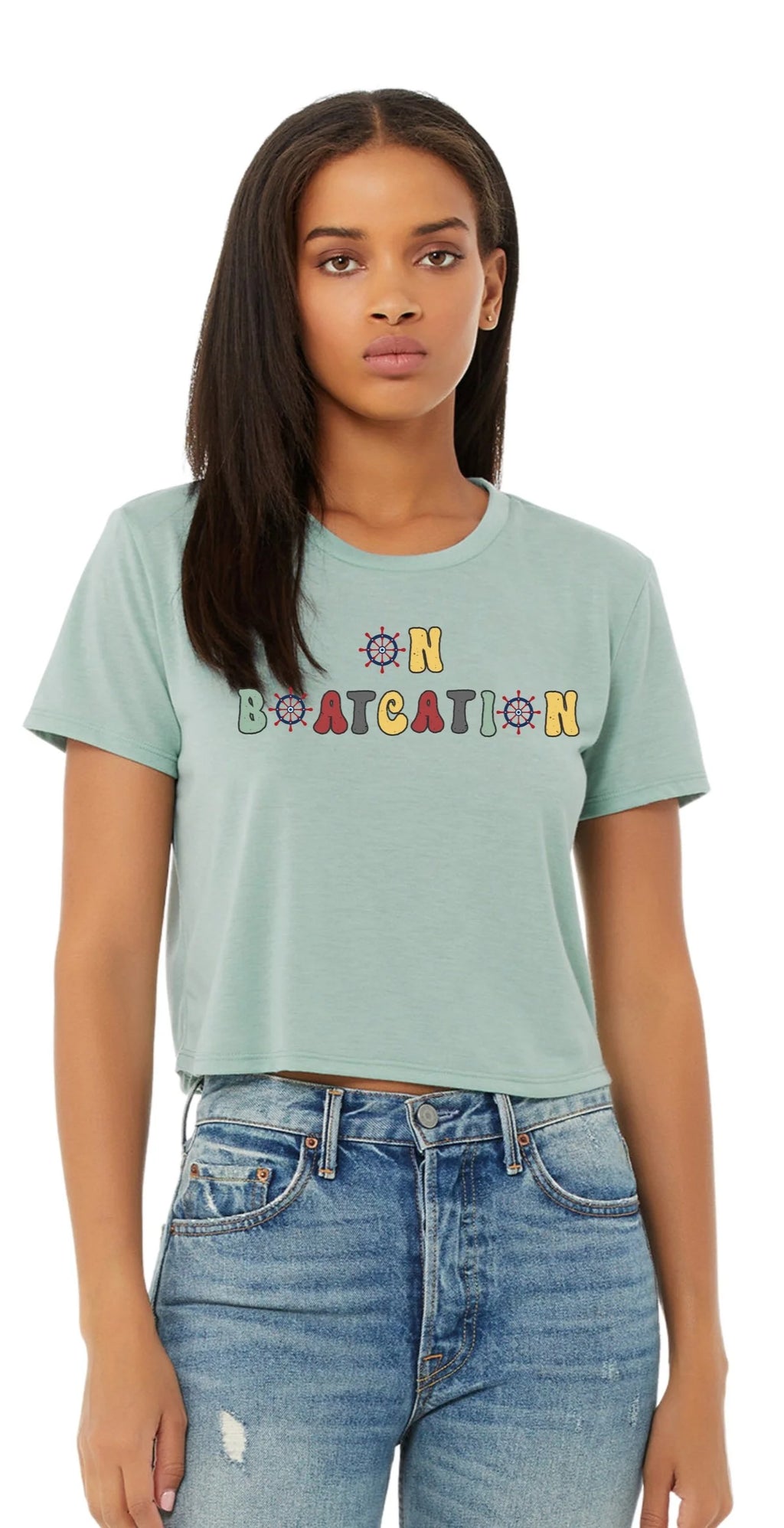 A woman modeling a light mint green cropped t-shirt with a colorful graphic text that reads 'ON BOATCATION' in yellow, orange, and blue letters, paired with blue jeans against a plain white background.