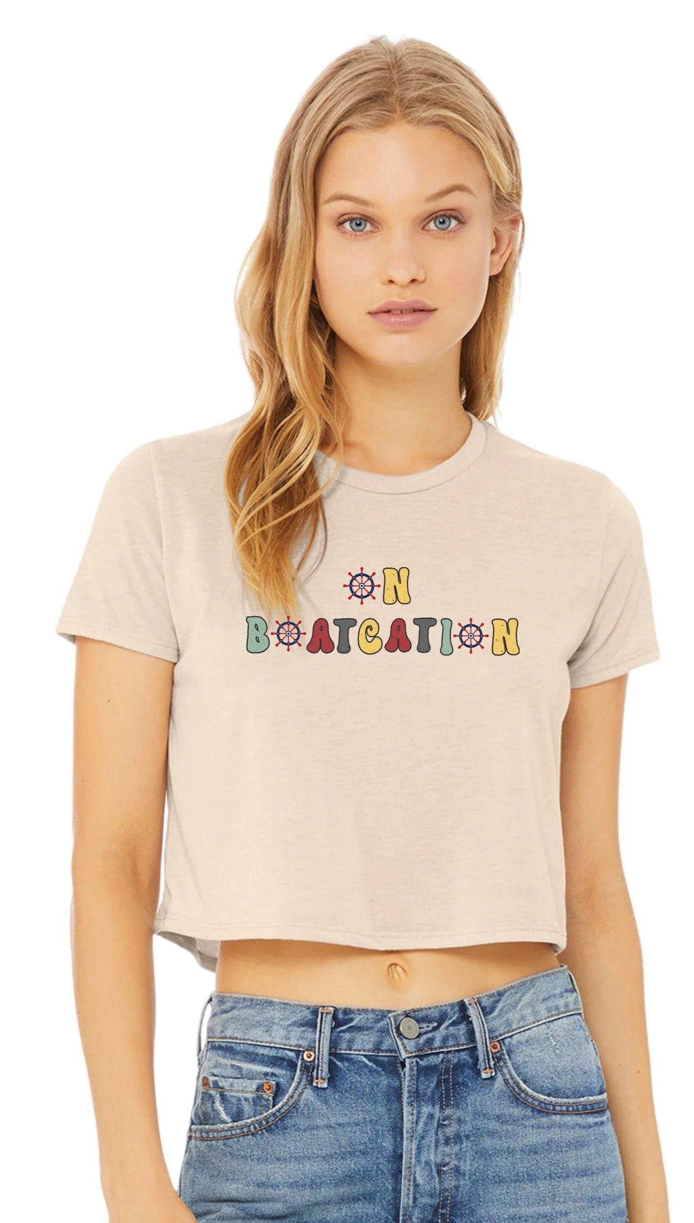 A blonde woman wearing a beige cropped short-sleeve t-shirt with a colorful retro-style graphic that reads 'ON BOATCATION' across the chest, paired with blue jeans, against a plain white background.