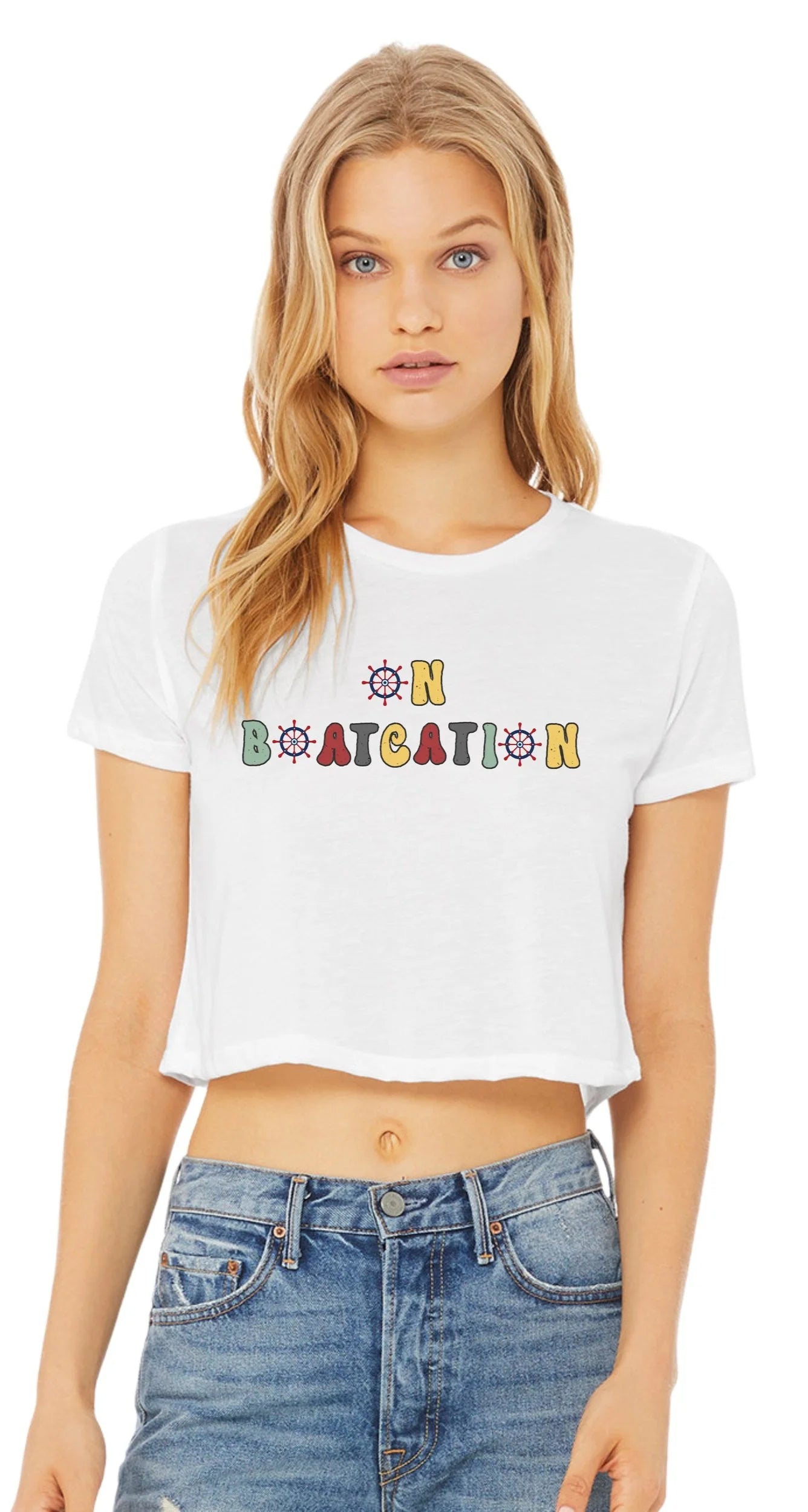 A blonde woman wearing a white cropped short-sleeve t-shirt with a colorful retro-style graphic that reads 'ON BOATCATION' across the chest, paired with blue jeans, against a plain white background.