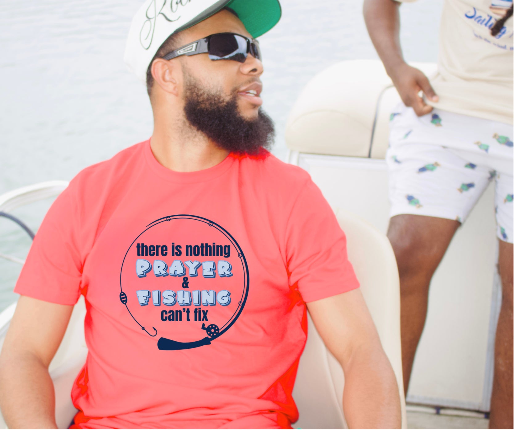 A man wearing a red t-shirt with a graphic that reads 'there is nothing PRAYER & FISHING can fix' in blue and black text, sitting on a boat. He is wearing a green and white cap and sunglasses, with another person partially visible in the background.