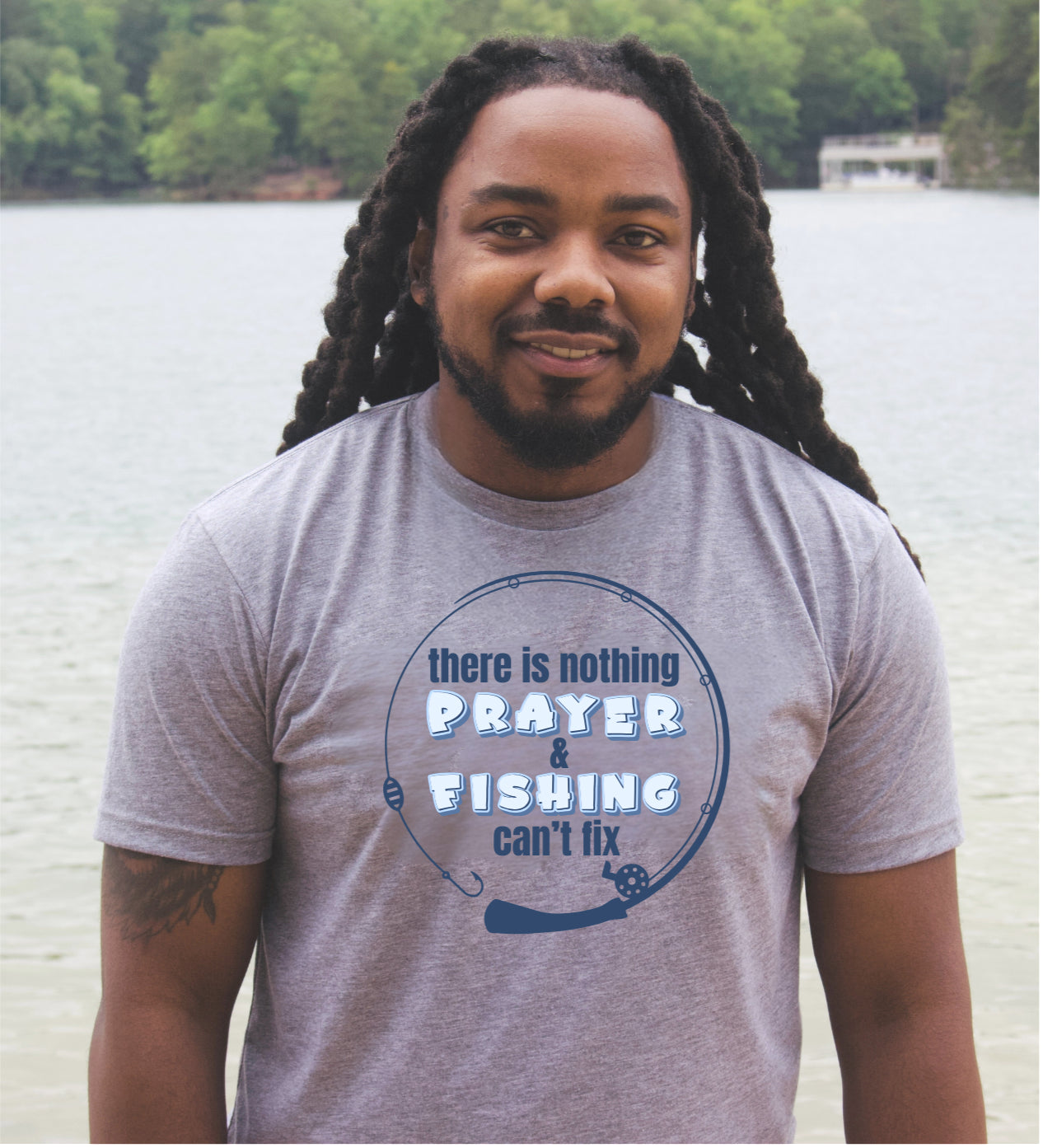 A man with long black locks stands outdoors by a lake wearing a heather gray t-shirt with a blue fishing-themed graphic that reads 'there is nothing PRAYER FISHING can't fix'.