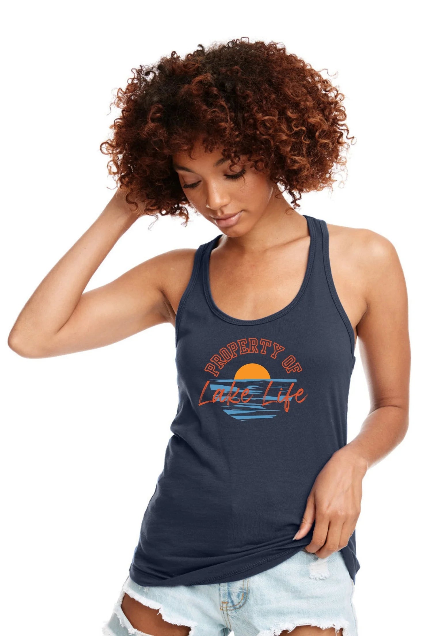 A woman with curly brown hair wearing a navy blue racerback tank top with a retro sunset graphic that reads 'Property of Lake Life' in orange and light blue, paired with light-wash denim shorts, against a plain white background.