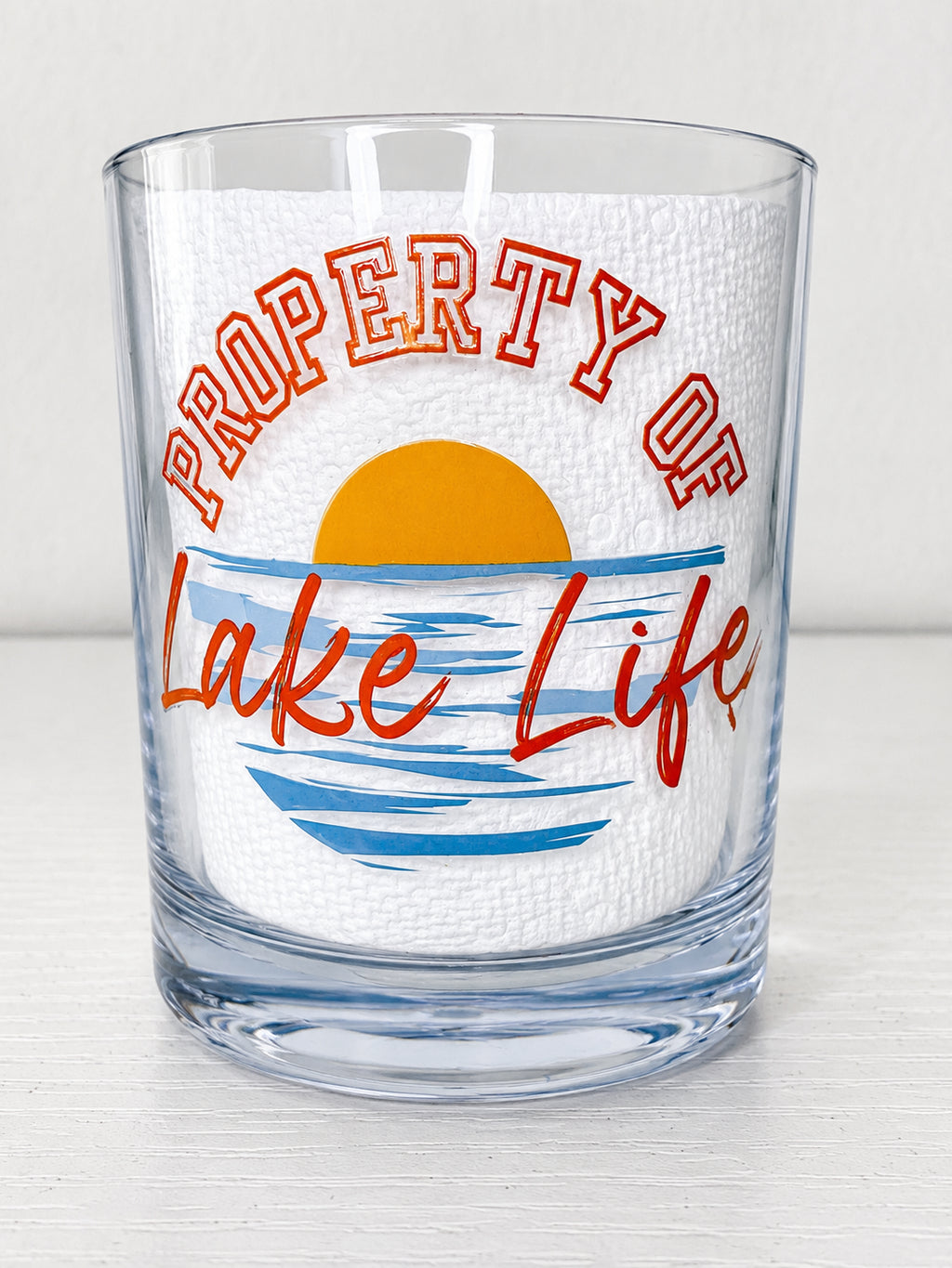 A clear rocks glass featuring a printed design that reads 'PROPERTY OF Lake Life' with a yellow sun and blue water graphic on a white textured insert, set against a light gray background.