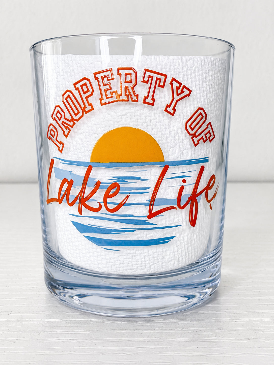 A clear rocks glass featuring a printed design that reads 'PROPERTY OF Lake Life' with a yellow sun and blue water graphic on a white textured insert, set against a light gray background.