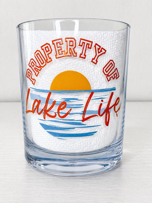 A clear rocks glass featuring a printed design that reads 'PROPERTY OF Lake Life' with a yellow sun and blue water graphic on a white textured insert, set against a light gray background.