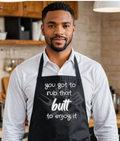 A man wearing a black kitchen apron with white text that reads 'you got to rub that butt to enjoy it', standing in a warm, rustic kitchen environment.