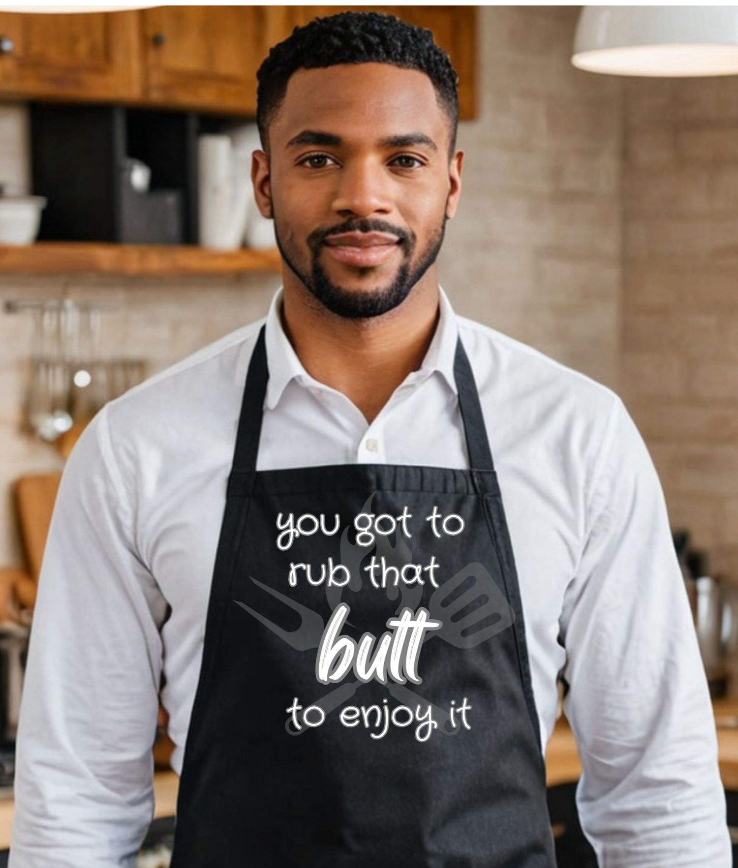A man wearing a black kitchen apron with white text that reads 'you got to rub that butt to enjoy it', standing in a warm, rustic kitchen environment.