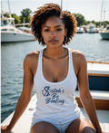 A woman with curly brown hair sits on a boat wearing a white tank top with the text 'Sistah's Be Boating' in navy blue script. The background shows a marina with water, boats, and trees.