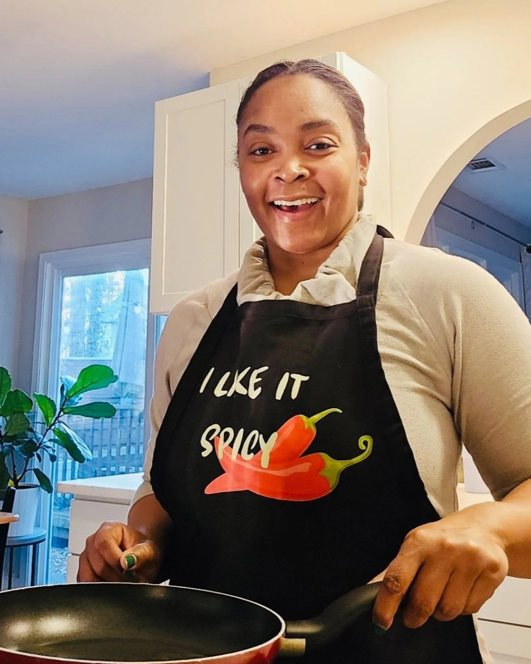 A woman smiling in a kitchen while wearing a black apron with the text 'I LIKE IT SPICY' and a red chili pepper graphic. She is holding a frying pan.