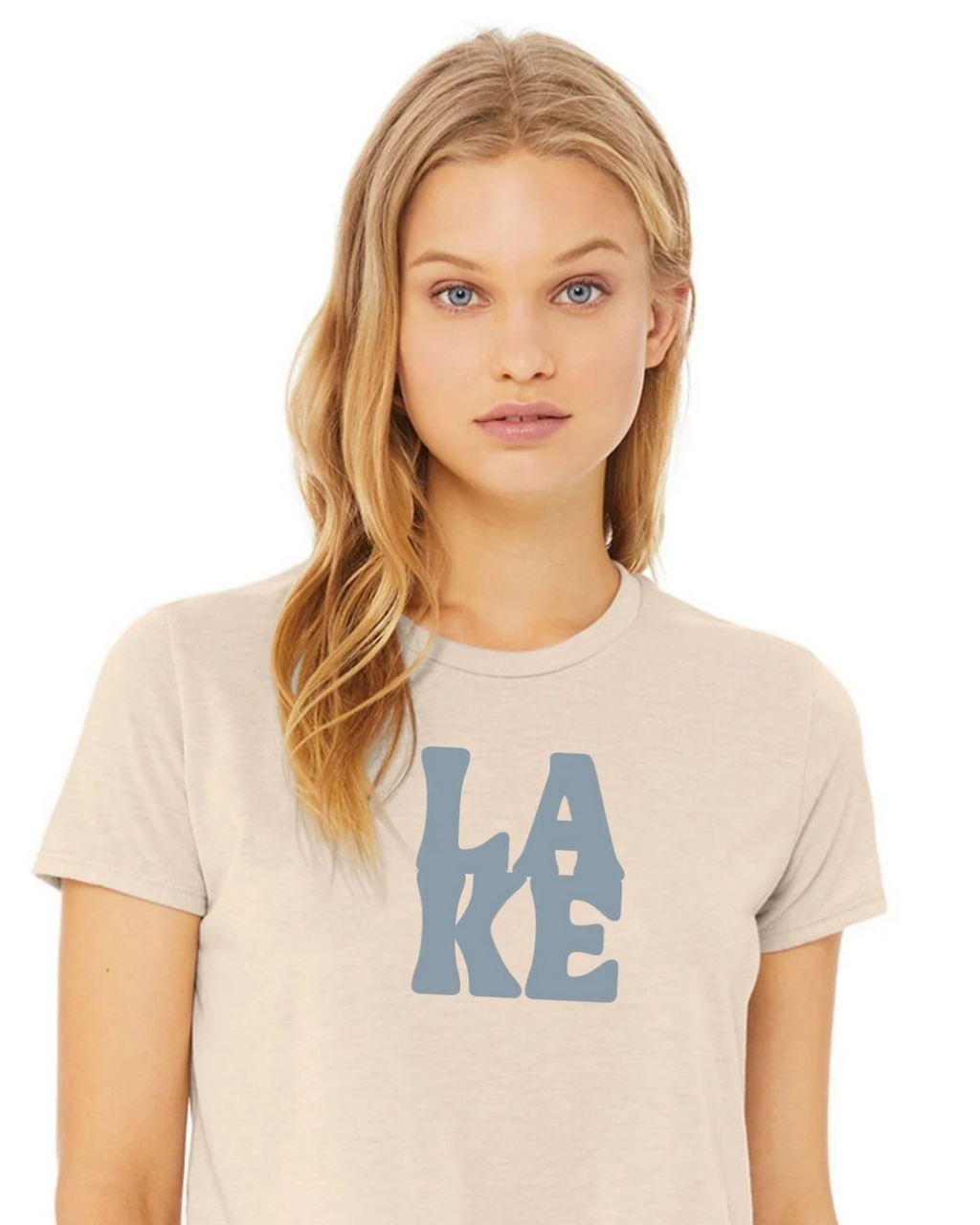 A woman with blonde hair wearing a light beige crewneck t-shirt with a blue graphic print spelling 'LAKE' on the front, set against a plain white background.