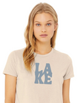 A woman with blonde hair wearing a light beige crewneck t-shirt with a blue graphic print spelling 'LAKE' on the front, set against a plain white background.