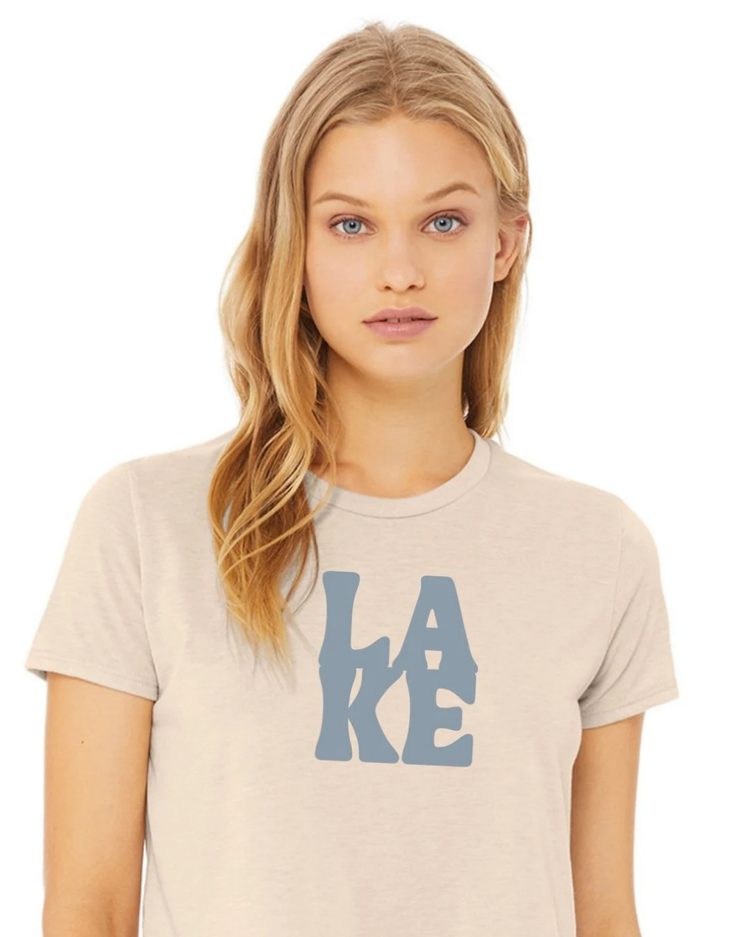 A woman with blonde hair wearing a light beige crewneck t-shirt with a blue graphic print spelling 'LAKE' on the front, set against a plain white background.