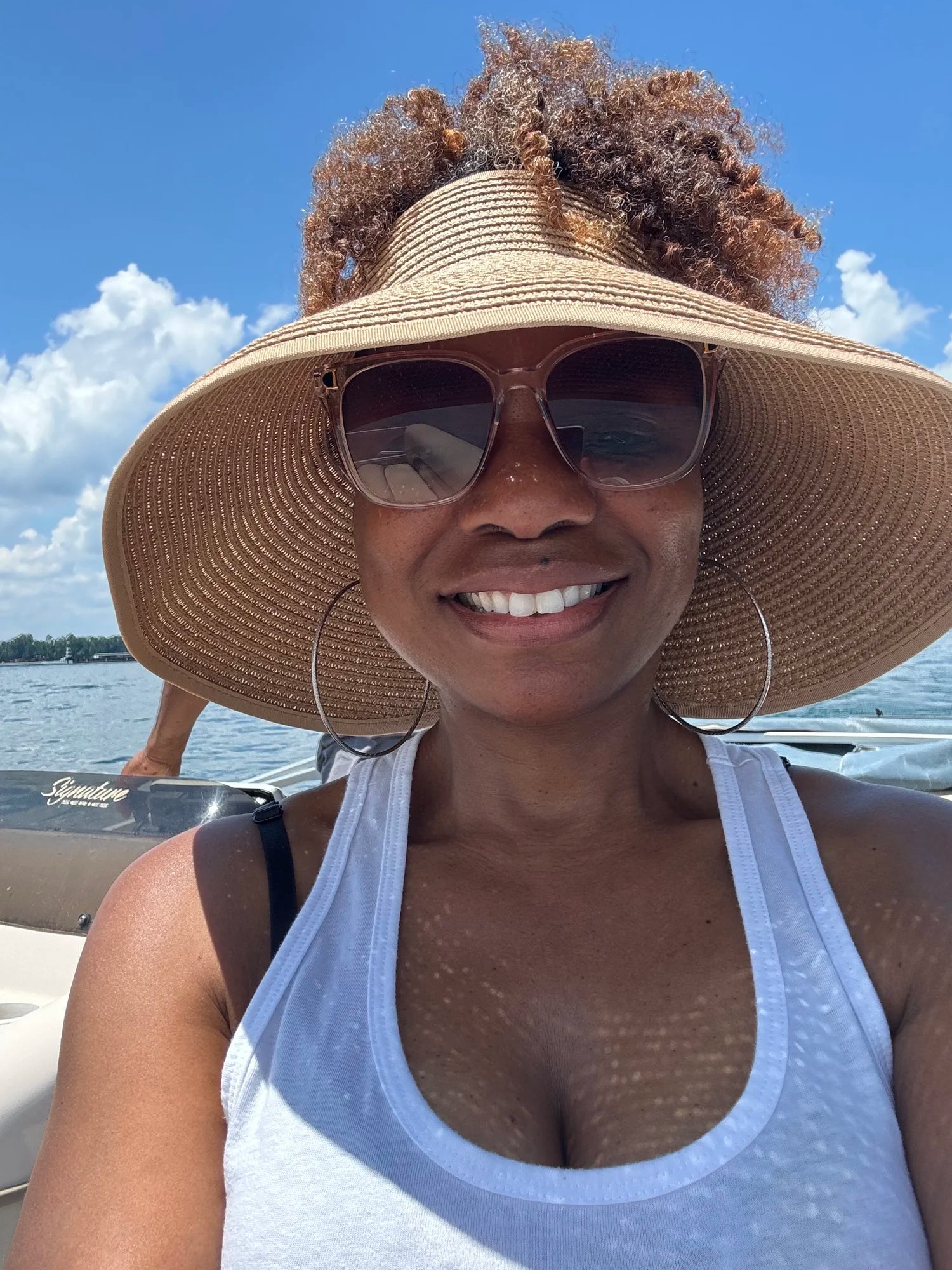 A woman on a boat wearing a large straw sun hat, sunglasses, hoop earrings, and a white tank top, with water and blue sky in the background.