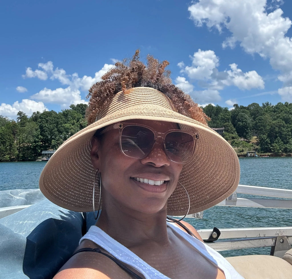 A woman wearing a large woven straw sun hat and sunglasses takes a selfie on a boat with water, trees, and a blue sky in the background.