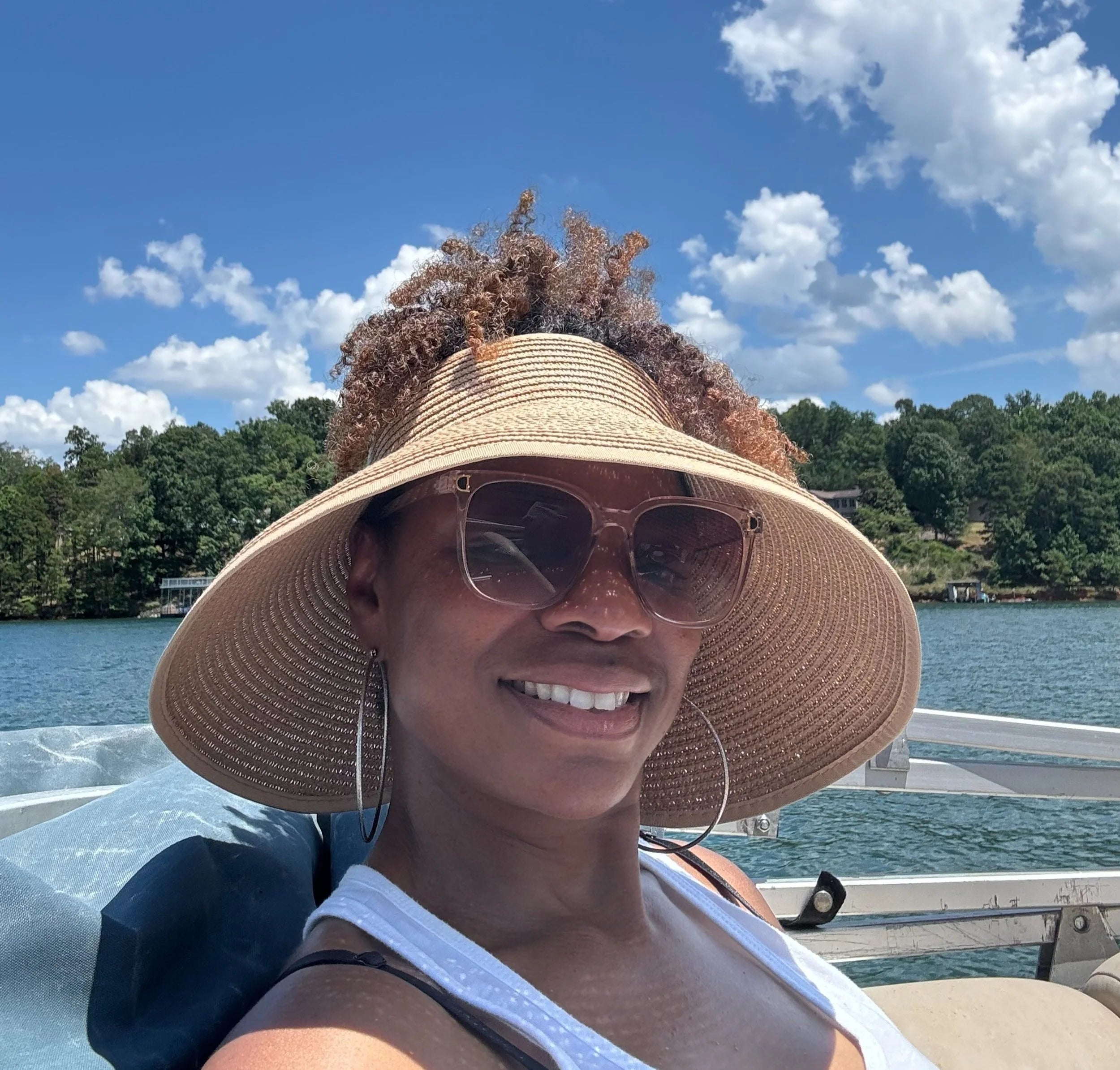 A woman wearing a large woven straw sun hat and sunglasses takes a selfie on a boat with water, trees, and a blue sky in the background.