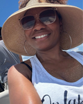 A smiling woman outdoors wearing a large woven straw sun hat, sunglasses, hoop earrings, and a white tank top under a blue sky.