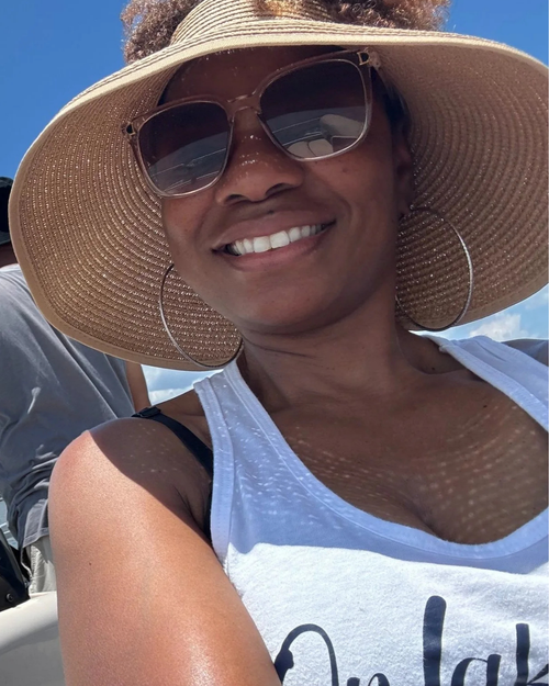 A smiling woman outdoors wearing a large woven straw sun hat, sunglasses, hoop earrings, and a white tank top under a blue sky.
