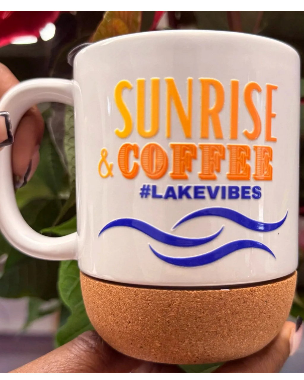 A white ceramic coffee mug with a cork base, held by a hand. The mug features the text 'SUNRISE & COFFEE #LAKEVIBES' in orange and blue lettering with blue wave graphics.