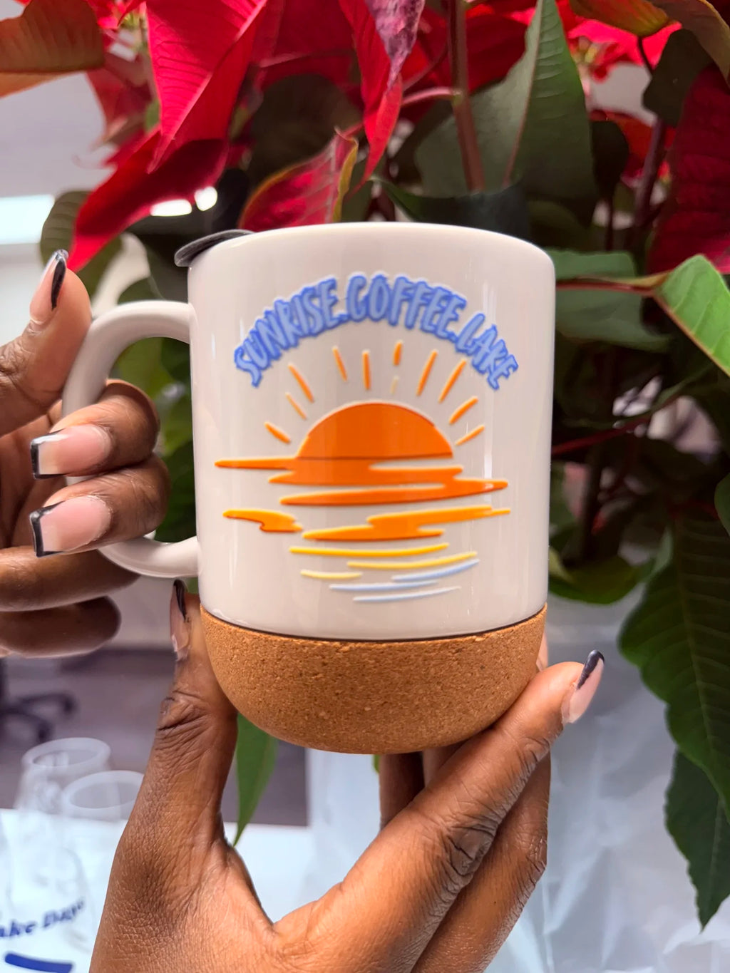 A person holds a ceramic coffee mug with a cork base. The mug is cream-colored with a blue and orange sunset graphic and the text 'SUNRISE COFFEE LAKE'.