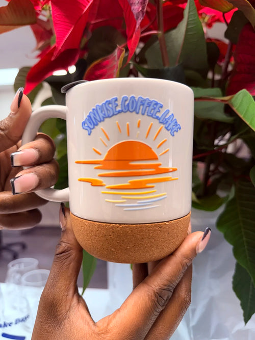 A person holds a ceramic coffee mug with a cork base. The mug is cream-colored with a blue and orange sunset graphic and the text 'SUNRISE COFFEE LAKE'.