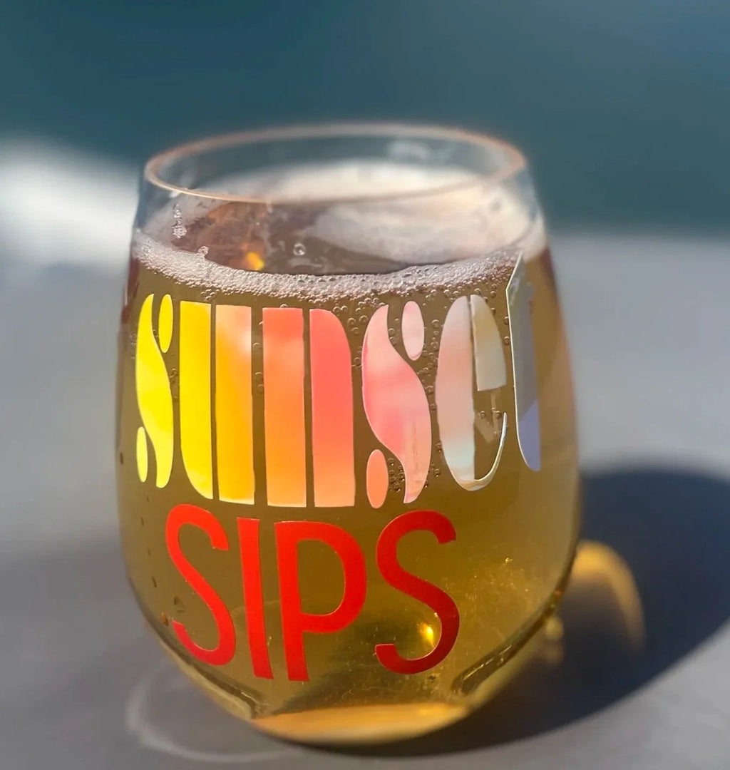 A shatterproof stemless wine glass filled with a golden beverage and foam, printed with the words 'SUNSET SIPS' in large retro lettering, photographed outdoors in bright sunlight.