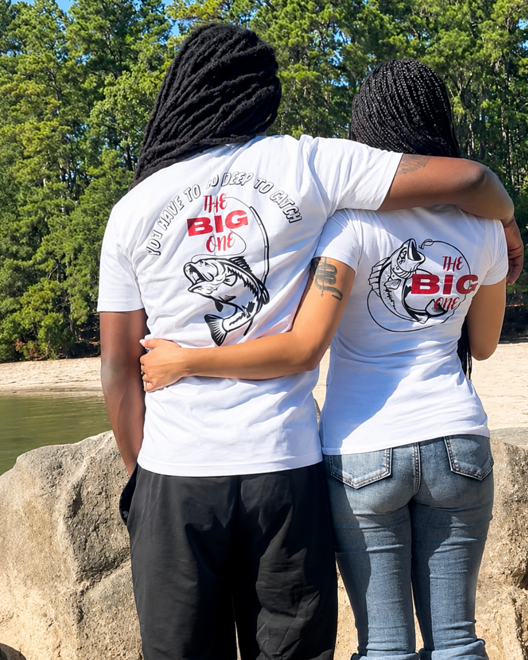 Two people standing outdoors by a lake with trees in the background, wearing matching white T-shirts with a large fish graphic and the text 'the BIG one' on the back.