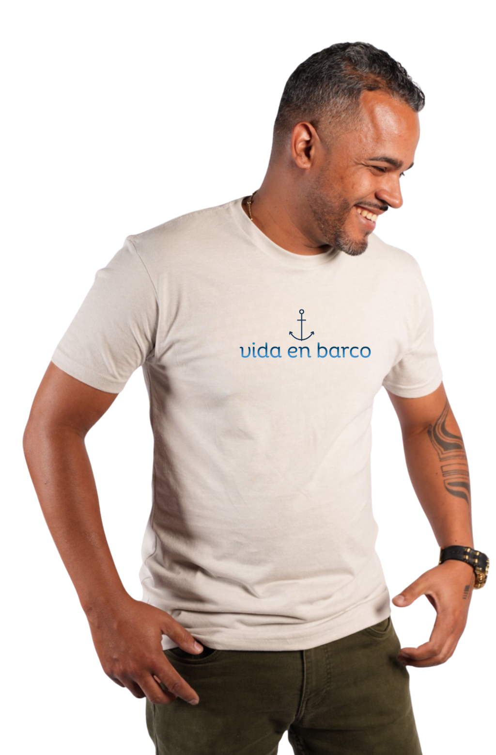 A man wearing a light beige crewneck t-shirt with a blue anchor graphic and the text 'vida en barco' printed on the chest, paired with olive green pants, set against a plain white background.